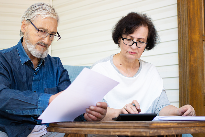 Serious focused mature couple reading paper documents and calculating bills to pay or checking domestic finances, senior family managing, planning budget, expenses, sitting on sofa at home terrace